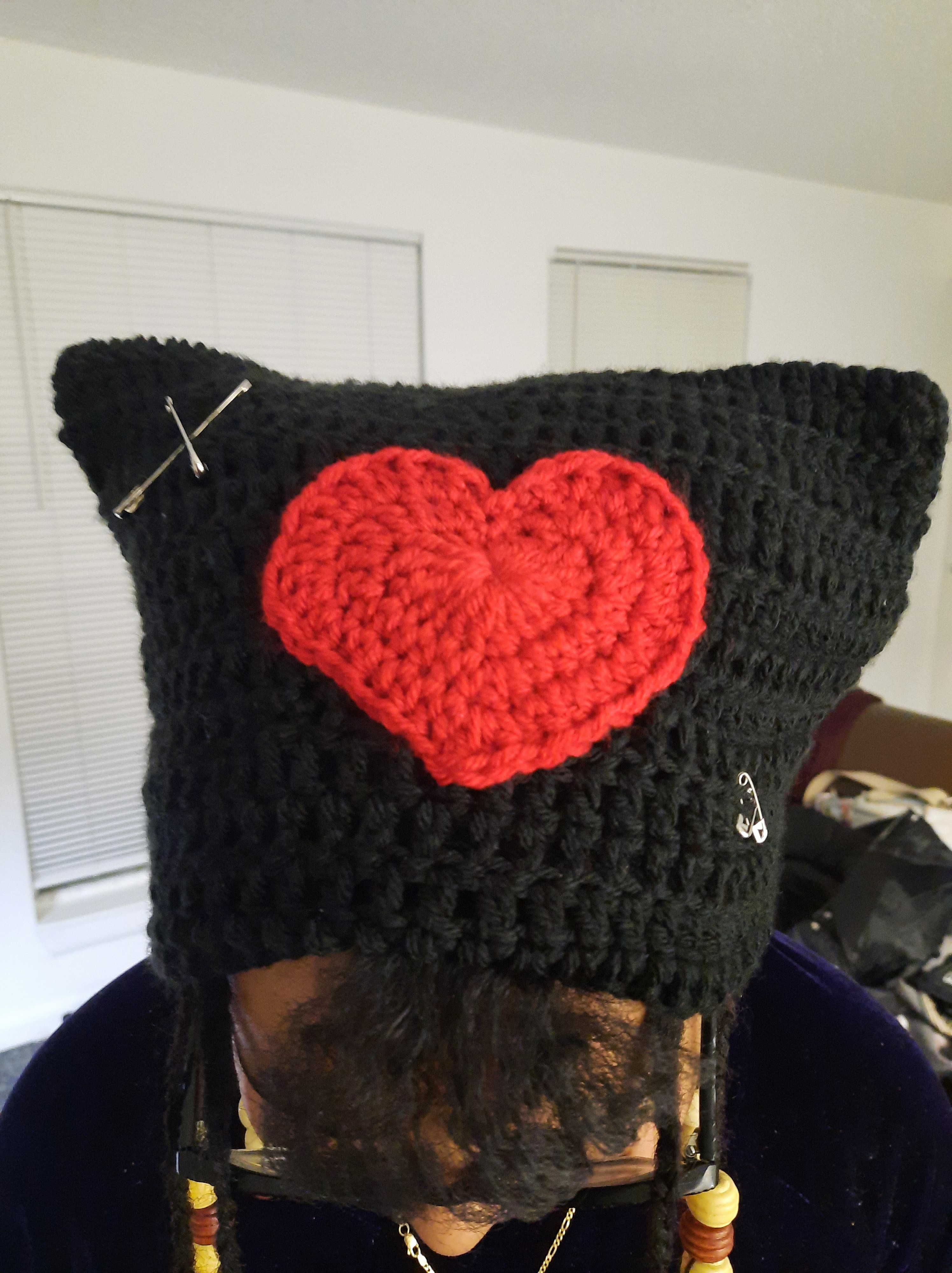 Black crochet cat hat with a red crochet heart in the middle and safety pins on the corners