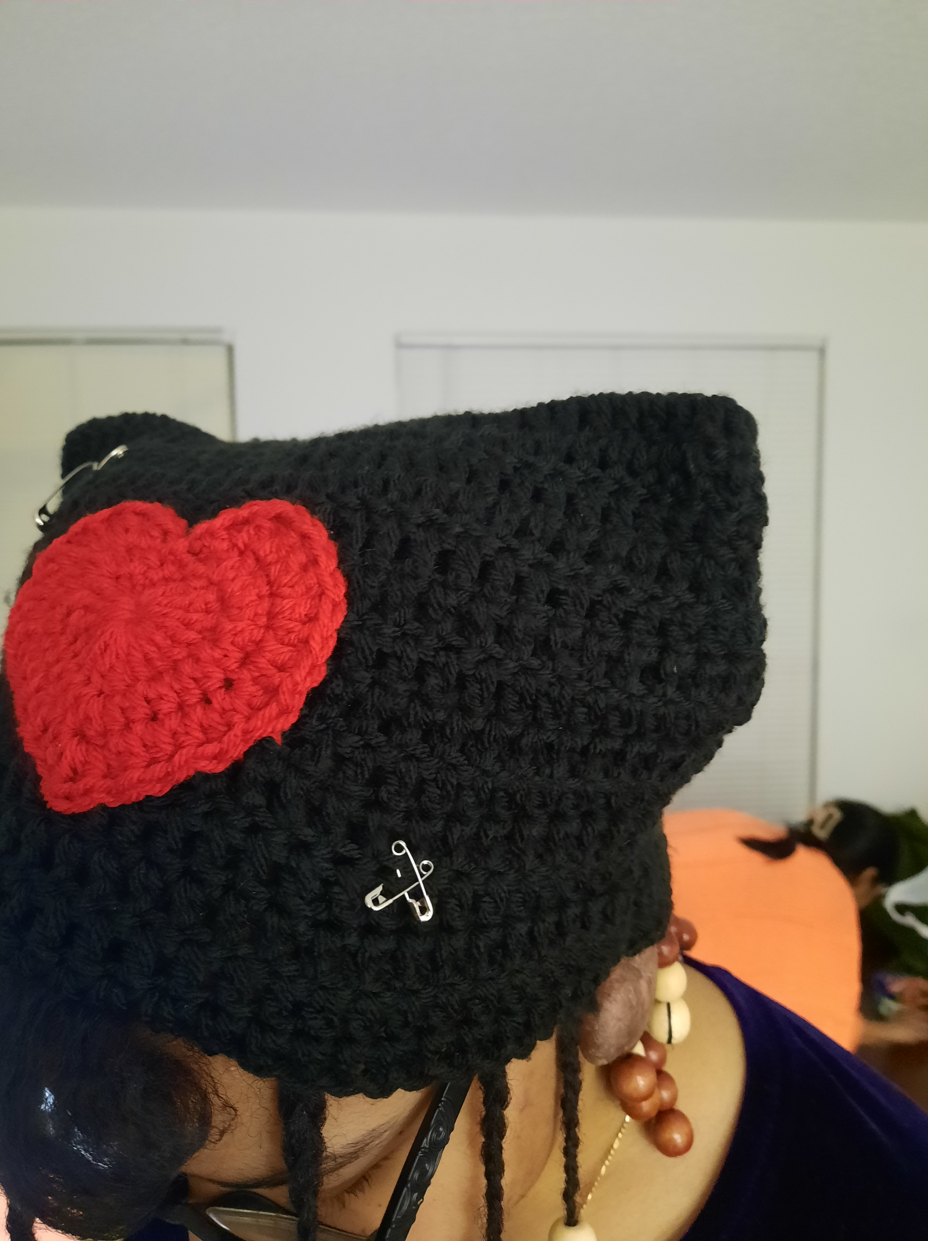 Black crochet cat hat with a red crochet heart in the middle and safety pins on the corners