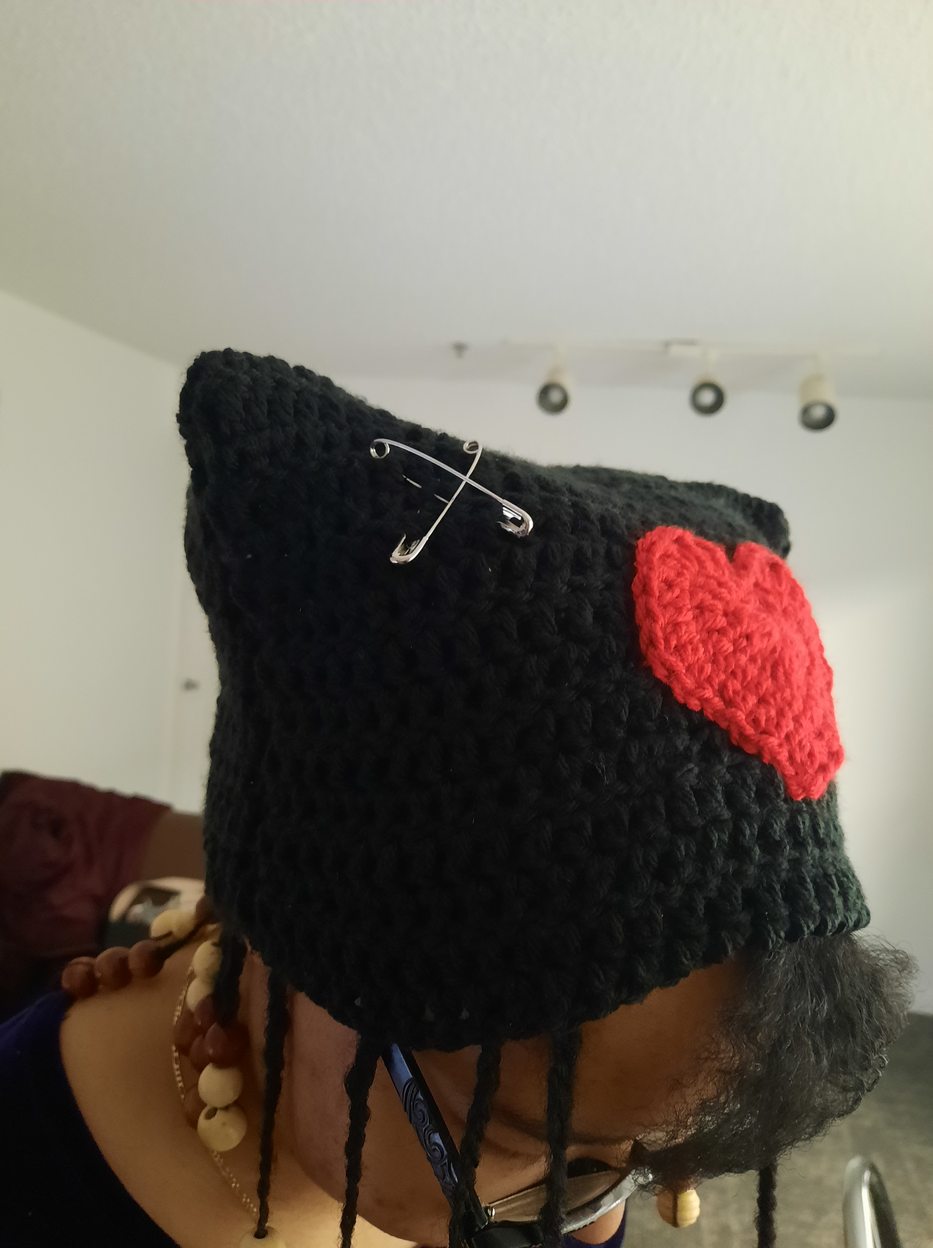 Black crochet cat hat with a red crochet heart in the middle and safety pins on the corners